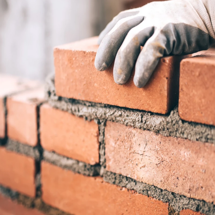 Hand wearing a grey glove placing a red brick on a wall, the mortar has Feb Wintamix Frost Proofer 5ltr added so it can be used in colder conditions.