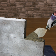 An image of a person using a trowel to apply mortar with Feb Wintamix Frost Proofer 5ltr added between concrete blocks against a stone wall background.