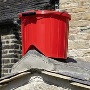 An image showing a roofers ridge bucket in Red with a black metal handle, mounted on a slate roof ridge