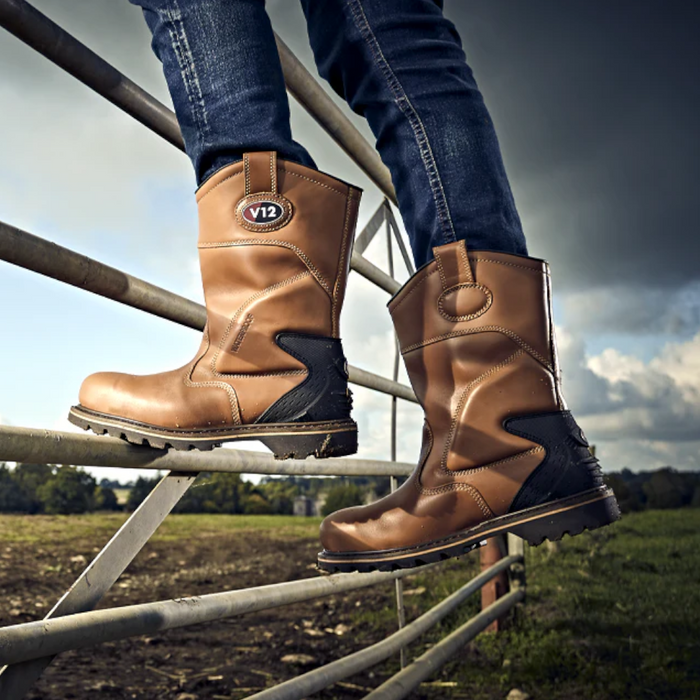 An image of someone wearing V12 Tomahawk V1250 Waterproof Rigger Boots climbing a metal gate to show they are versatile and for heavy use on all terrains. It is a light tan colour and has a black heel. The photo is taken on a muddy field.